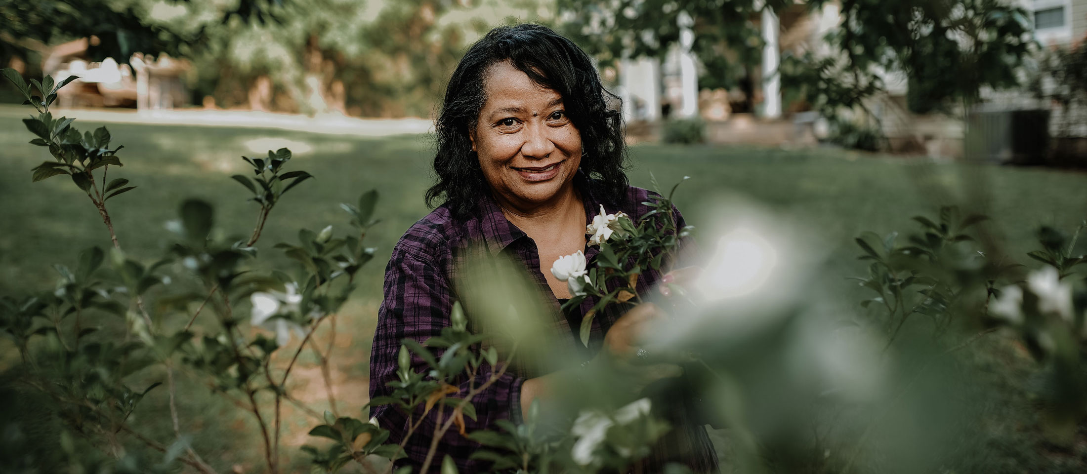 Woman peeking through a green plant, smiling with dark brown curly hair.